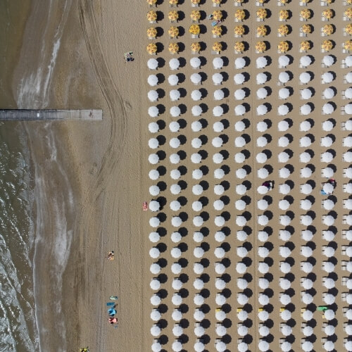 Vista aerea di una spiaggia affollata con ombrelloni bianchi, sdraio gialle e persone sparse sulla sabbia scura.