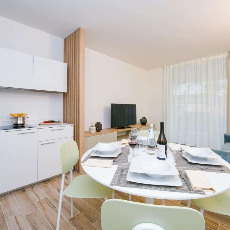 Salle à manger moderne avec table ronde pour quatre, cuisine à gauche et salon en arrière-plan, décor minimaliste.