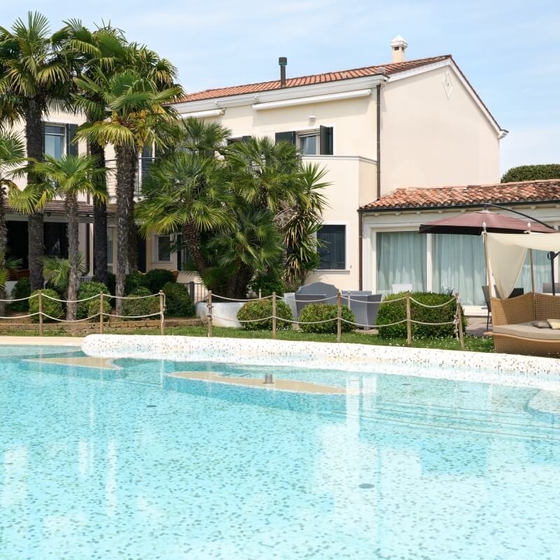 Scène paisible d'une piscine devant une maison méditerranéenne avec des chaises longues sous un parasol.