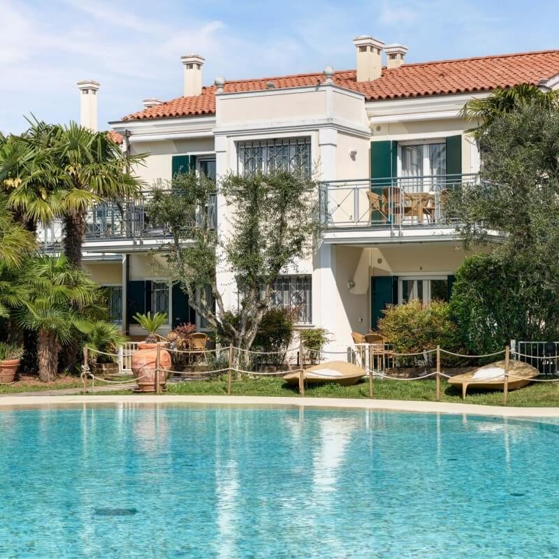 Une maison à deux étages avec un balcon, piscine bleue, chaises longues et jardin, évoquant la détente et le luxe.