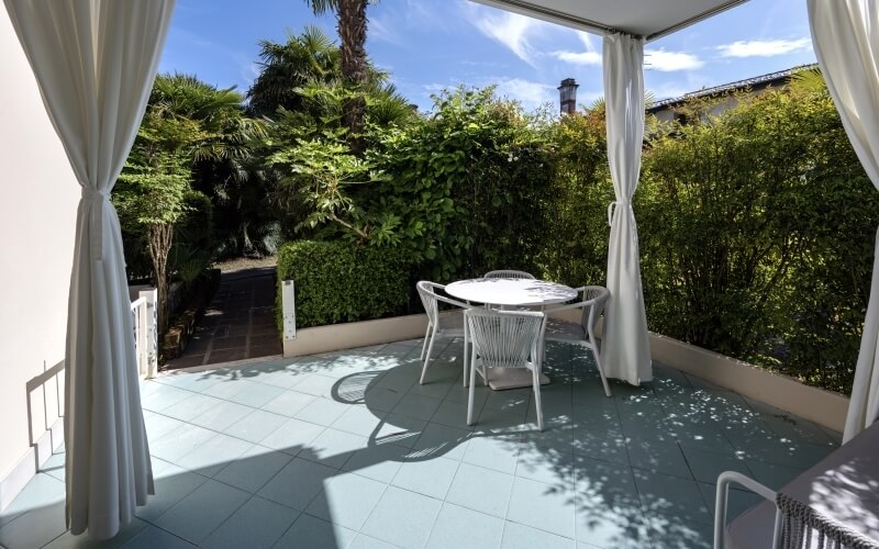 Ein einladender Patio mit weißen Fliesen, Rattanmöbeln und grüner Umgebung unter einem blauen Himmel mit Wolken.