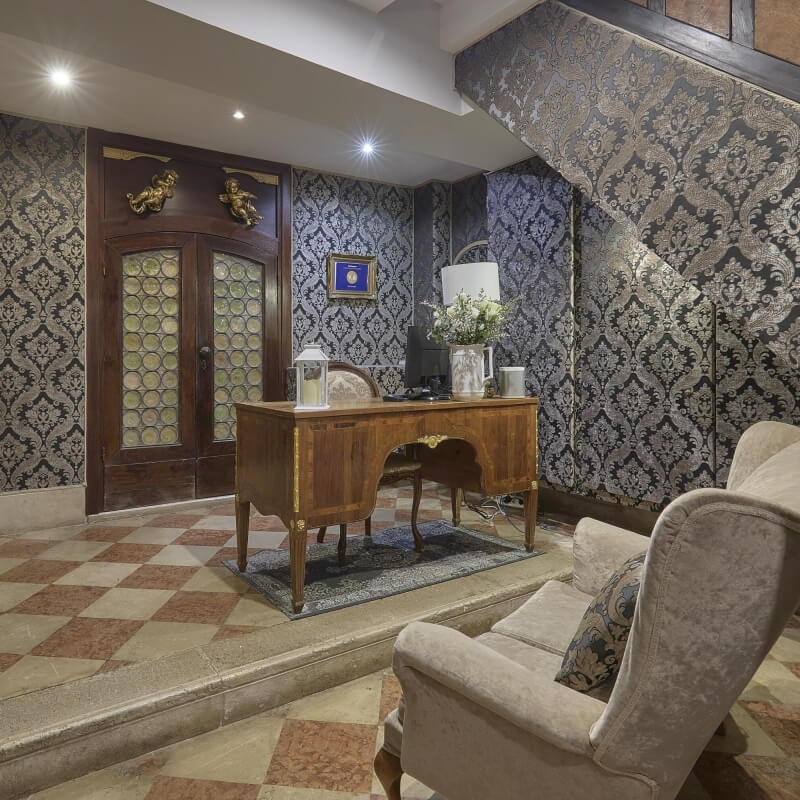 Sophisticated lobby with wooden desk, floral decor, blue cushions, and elegant wallpaper, exuding luxury and refinement.