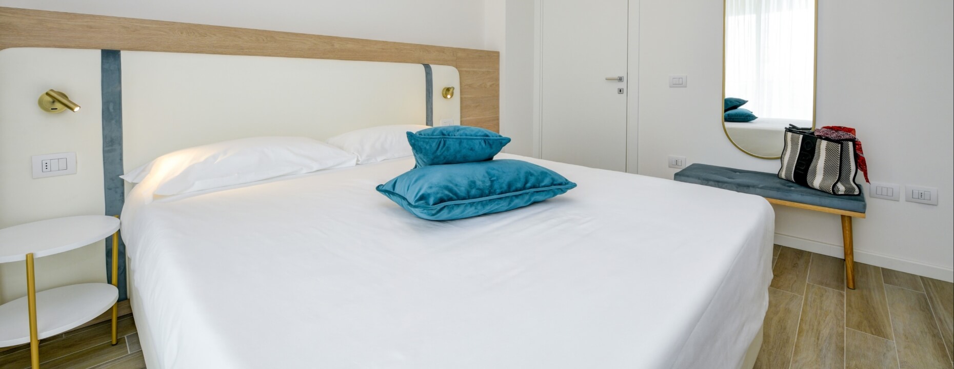 Camera d'hotel con letto bianco, cuscini teal e arredi eleganti.