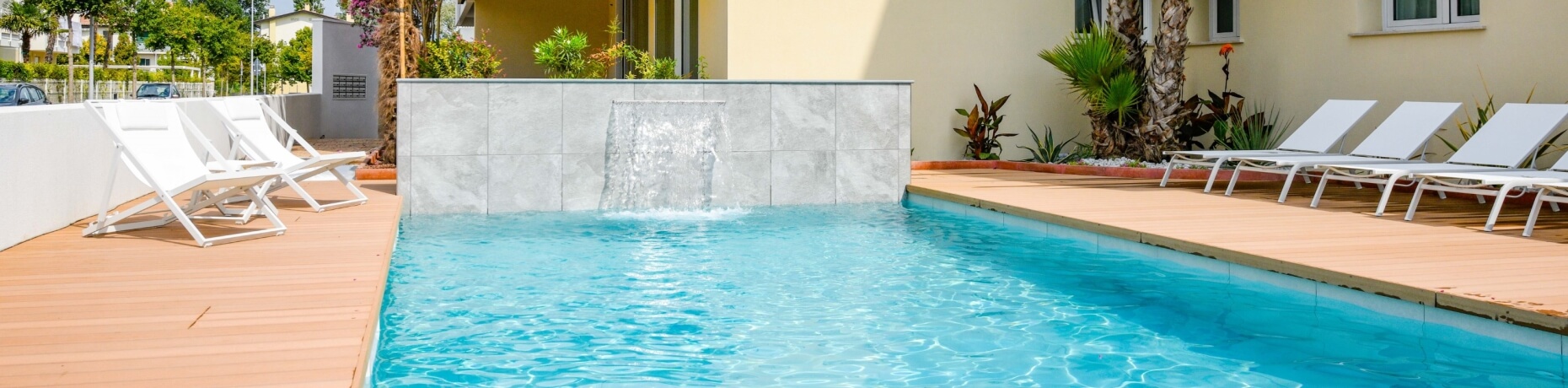 Scène de piscine sereine avec cascade, chaises longues blanches, plantes tropicales et mur de confidentialité en arrière-plan.