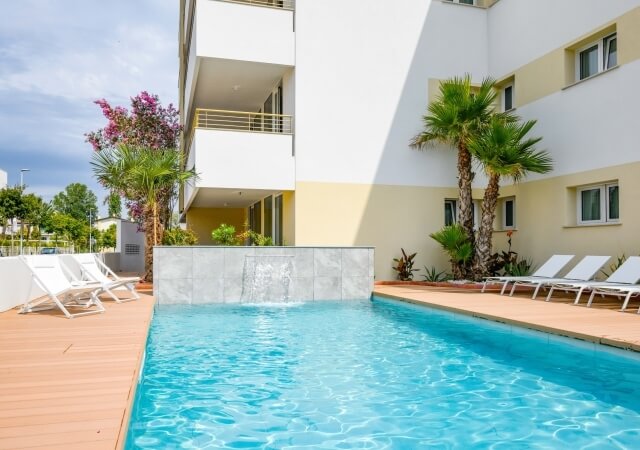 Piscina rettangolare turchese con cascata, circondata da un deck in legno e palme, con un moderno edificio sullo sfondo.