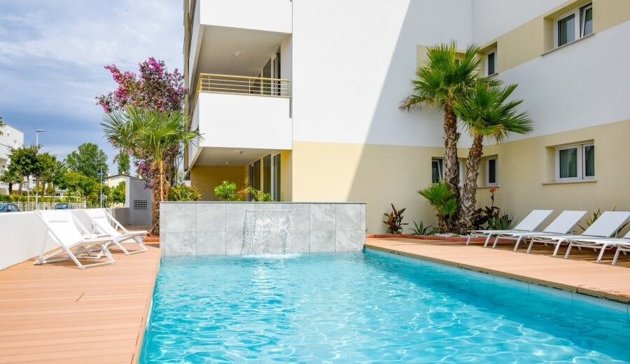 Una piscina circondata da un deck in legno, con un edificio bianco e giallo sullo sfondo e sdraio bianche a destra.