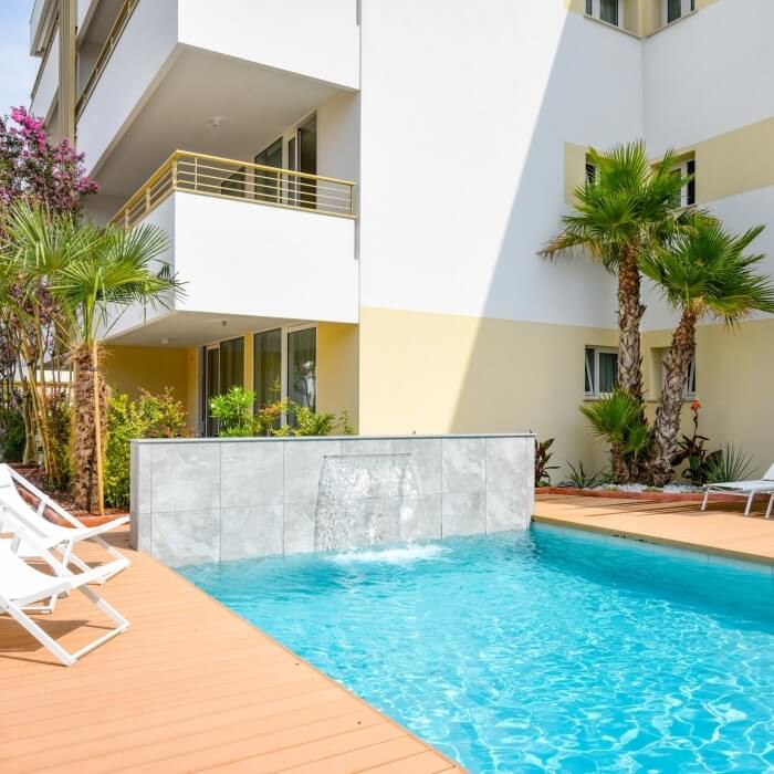 Una piscina serena con fontana, circondata da lettini e un edificio bianco e beige sullo sfondo, circondato da piante tropicali.