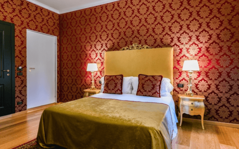 A stylish bedroom with a gold headboard bed, red pillows, nightstands with lamps, and patterned wallpaper.