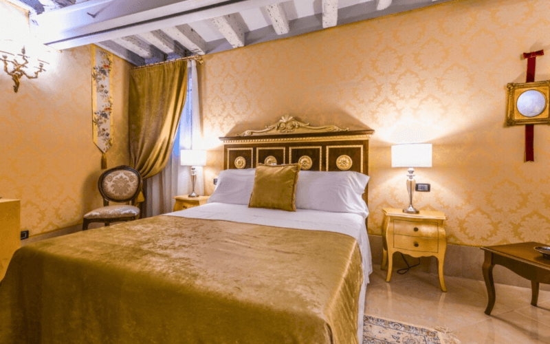 Schlafzimmer mit goldenen Akzenten, Bett mit weißer Bettdecke, Nachttische, Stuhl, Fenster mit gelben Vorhängen und Kronleuchter.