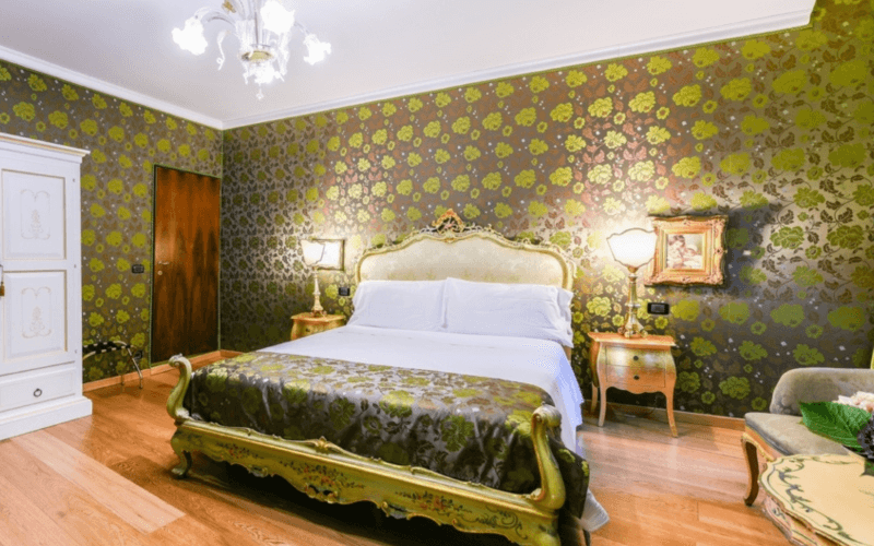 Helles Hotelzimmer mit Holzfußboden, grünem Bett, Schreibtisch, Stuhl, antiken Kronleuchtern und gemusterter Tapete.