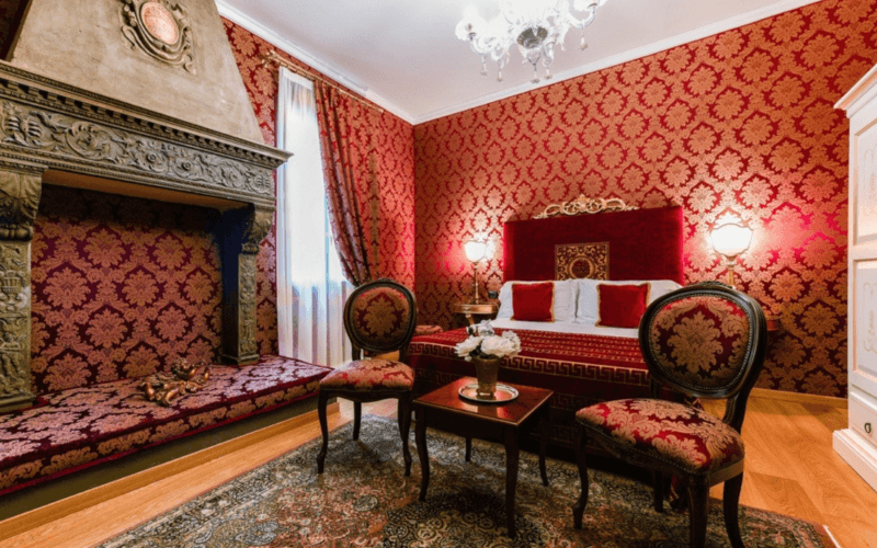 Lavish bedroom with red and gold decor, damask walls, ornate fireplace, crystal chandelier, and elegant furnishings.
