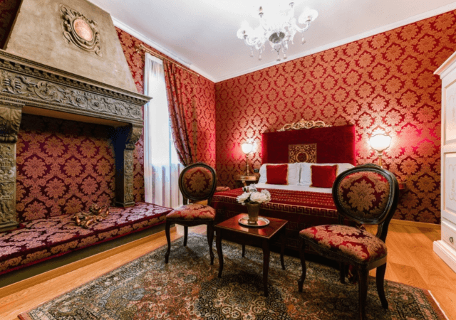 Chambre luxueuse avec motifs rouges et dorés, cheminée ornée, canapé plush, et lustre en cristal.