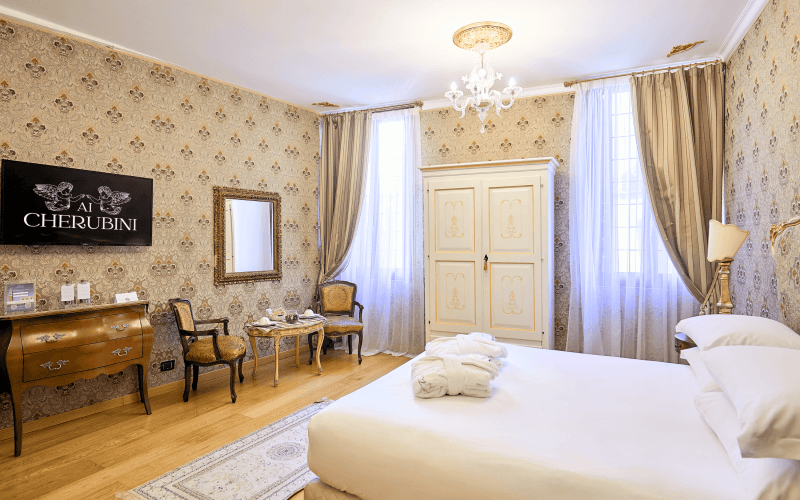A serene bedroom with a large bed, floral wallpaper, wooden floor, dresser, chairs, and a chandelier.