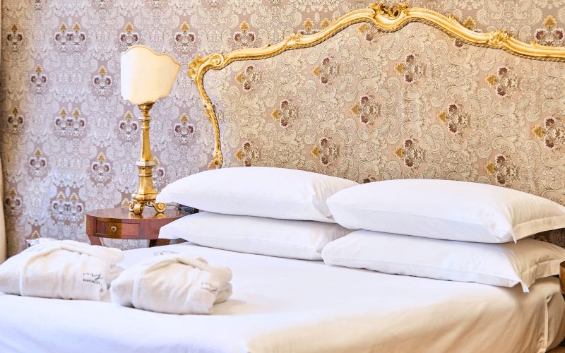 Luxuriöses Schlafzimmer mit goldener Kopfteil, weißer Bettdecke, Nachttischlampe und blumiger Tapete in Beige und Gold.