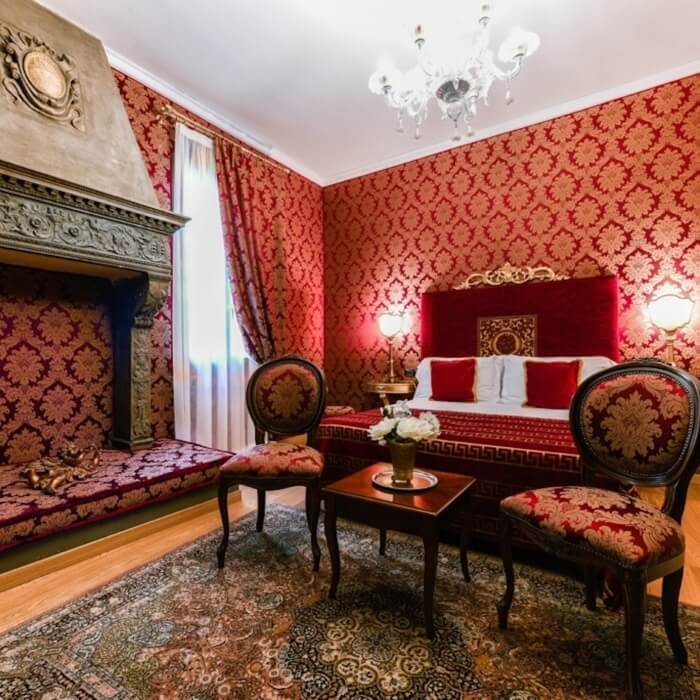 Camera da letto lussuosa con letto a baldacchino, tonalità rosso e oro, camino, e lampadario di cristallo.