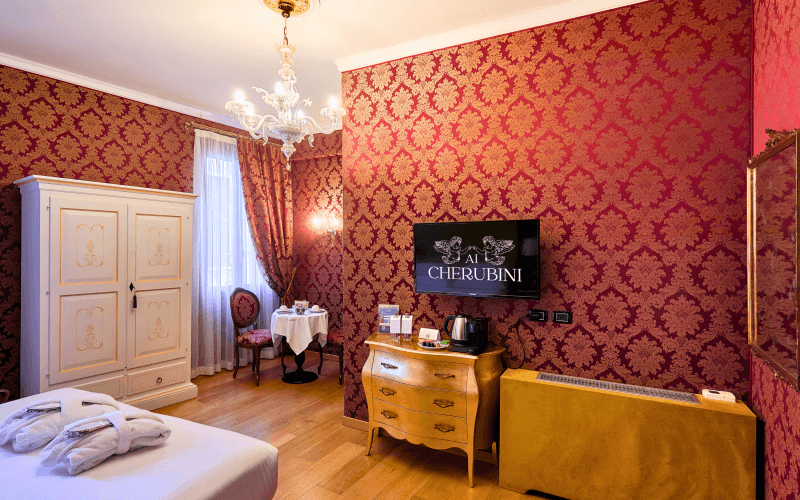 Luxurious hotel room with red and gold decor, white armoire, table and chairs, dresser with TV, and ornate patterns.