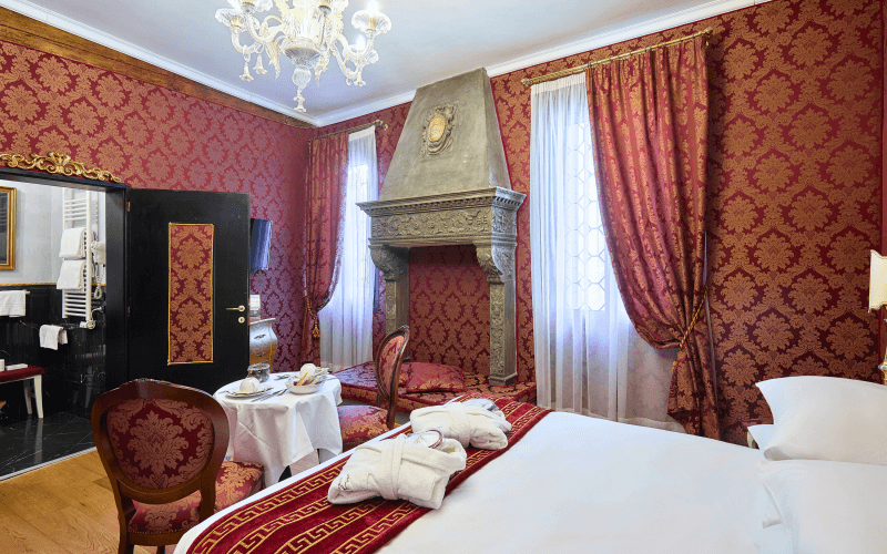 Lavish hotel room with red and gold wallpaper, king-size bed, fireplace, and breakfast table, featuring elegant decor.