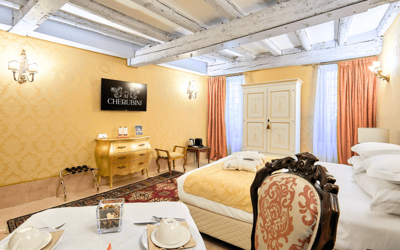 A bright hotel room with yellow and white decor, featuring a bed, desk, chair, dresser, and natural light from windows.