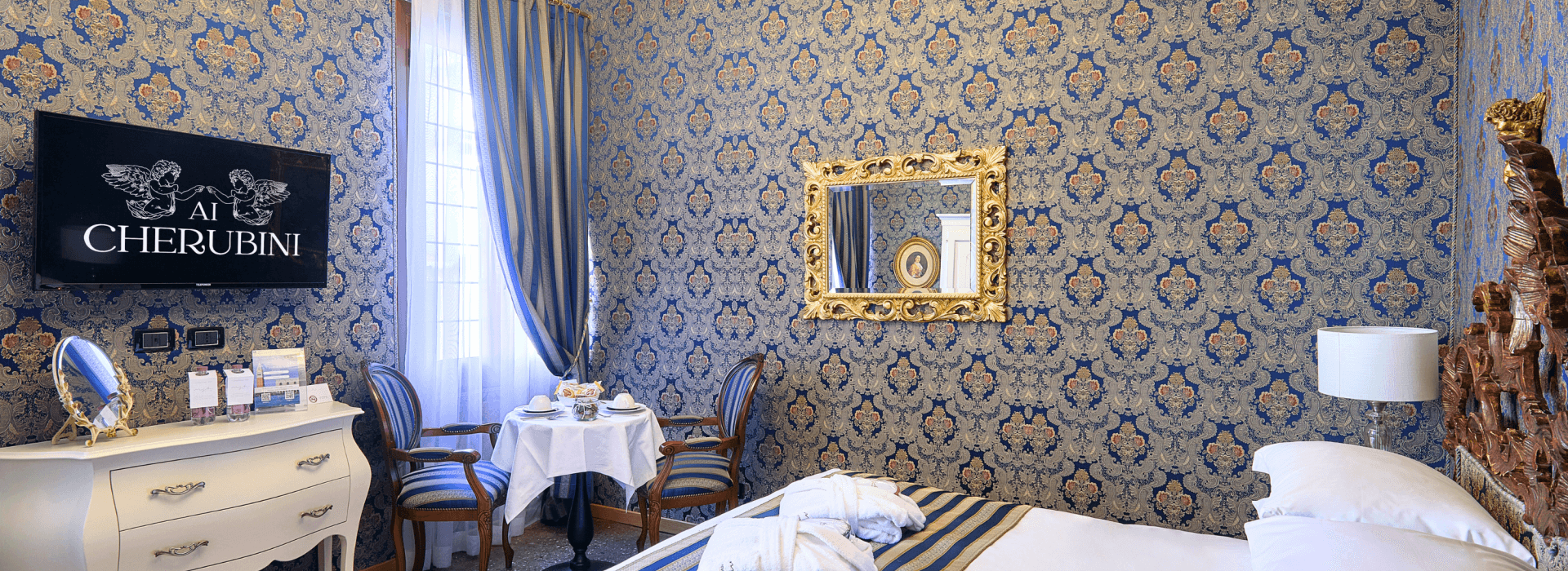 Chambre d'hôtel élégante avec papier peint bleu et or, lit double, commode blanche, table, miroir doré et vase de fleurs.
