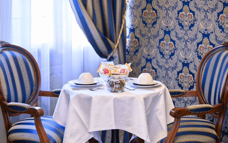Ein runder Tisch für zwei mit weißer Tischdecke, blauen und goldgestreiften Stühlen, Geschirr und Snacks in eleganter Umgebung.
