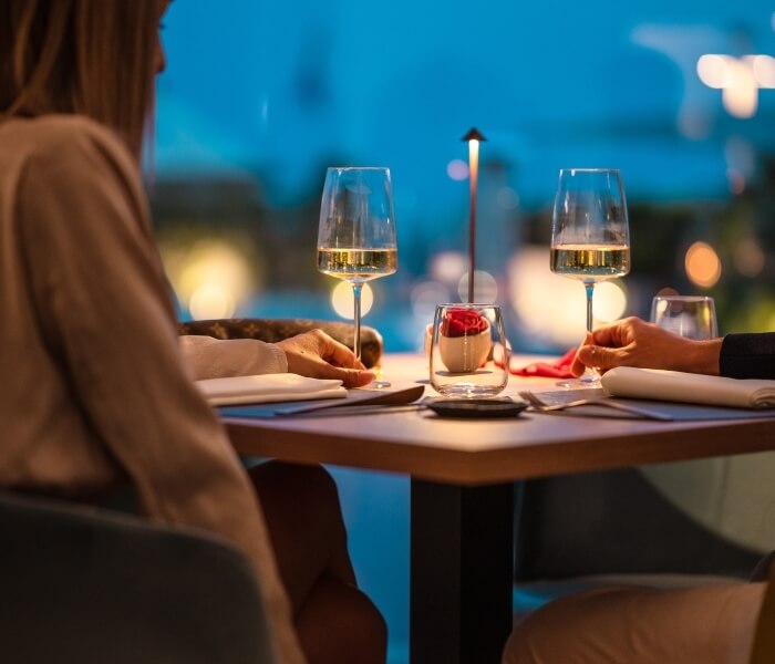 Cena romantica all'aperto con una coppia che brinda con vino bianco, circondata da una candela e una rosa galleggiante.