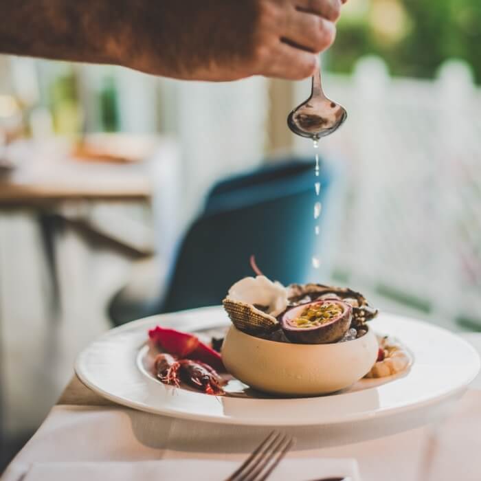 Una mano tiene un cucchiaio sopra un piatto bianco con ingredienti, in un ristorante sfocato sullo sfondo.