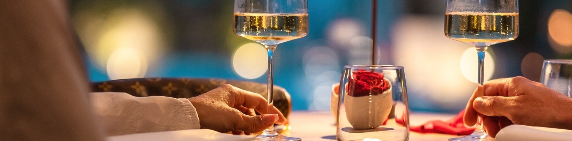 Due mani che tengono bicchieri di vino, con una rosa rossa e un tovagliolo rosso in un'atmosfera romantica.