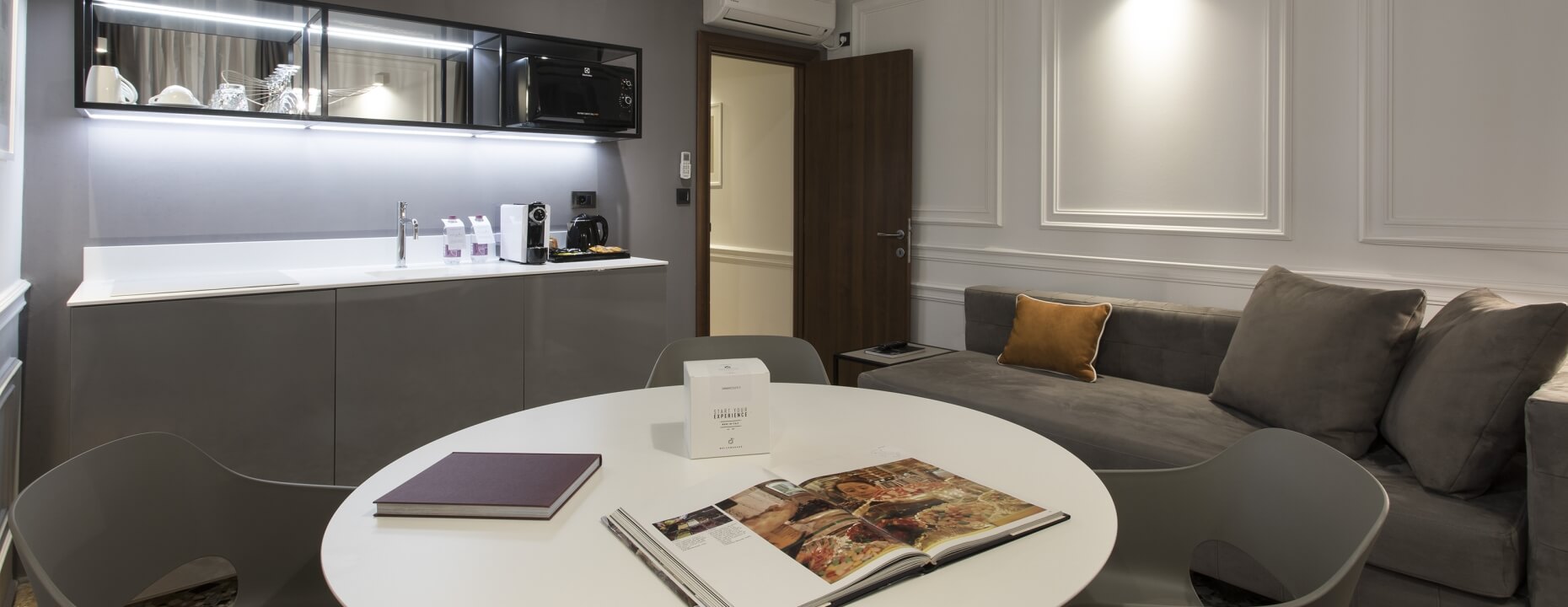 Suite d'hotel con divano grigio, tavolo rotondo e angolo cottura.