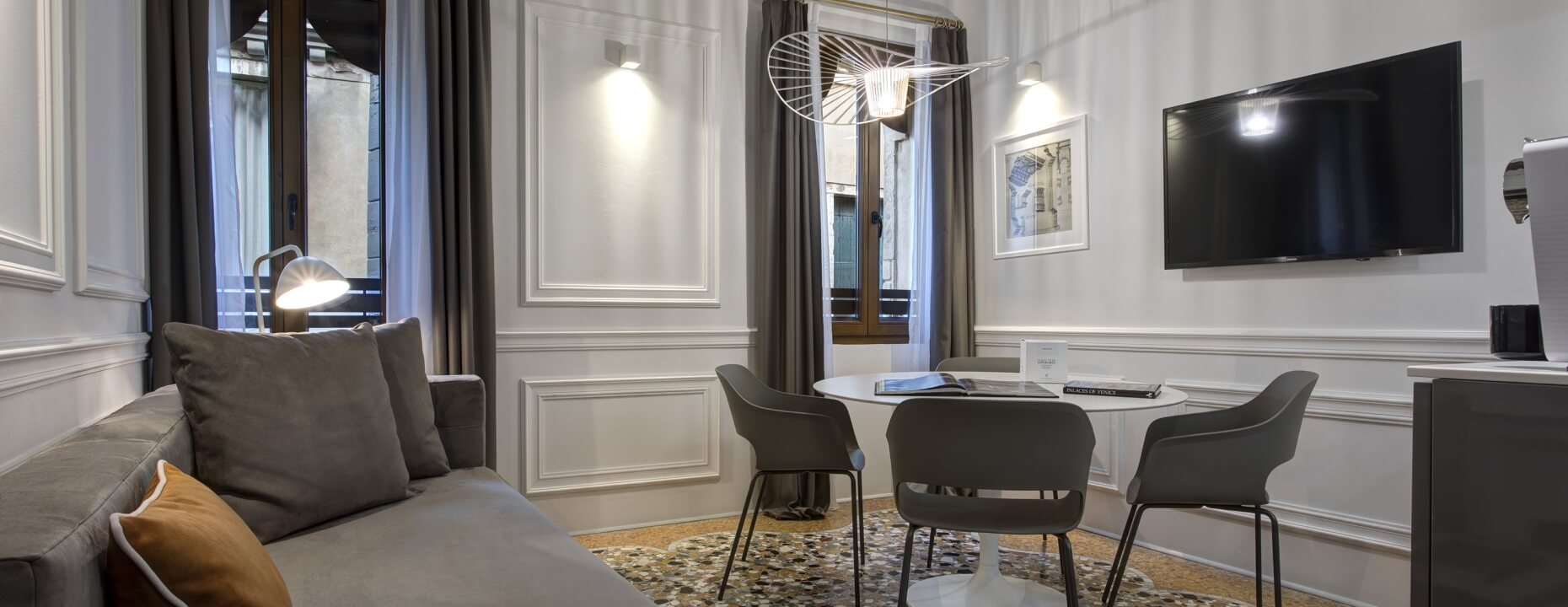 Camera d'hotel con divano grigio, tavolo bianco e TV.