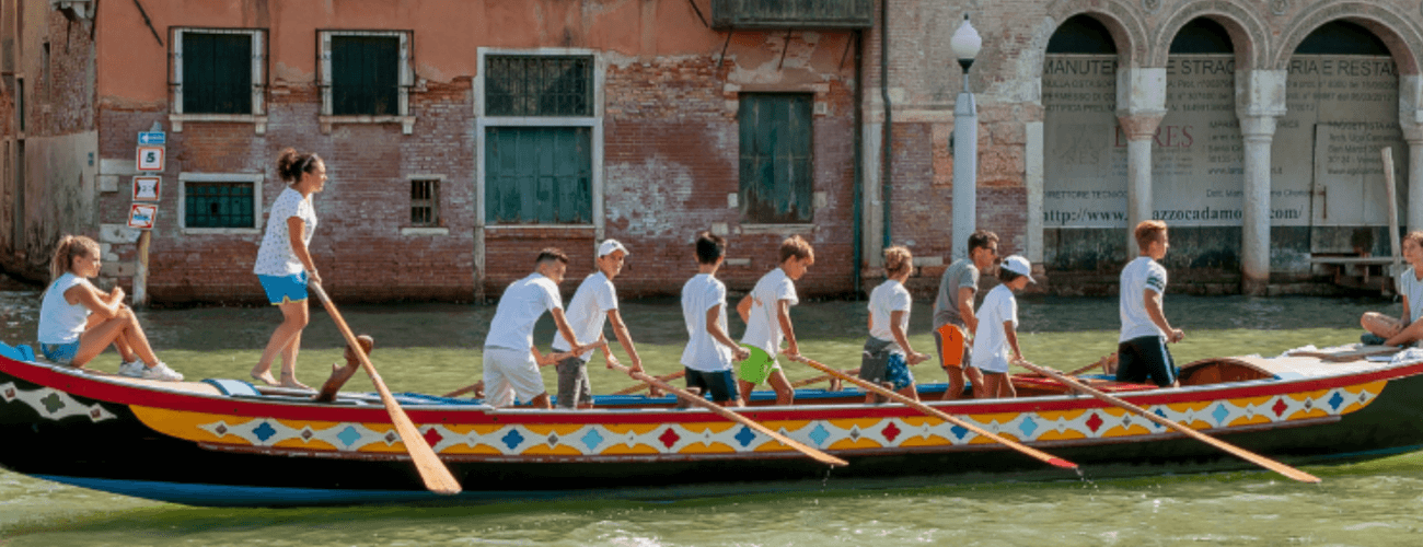 Barca colorata con giovani che remano su acqua verde, con edifici storici e moderni sullo sfondo.