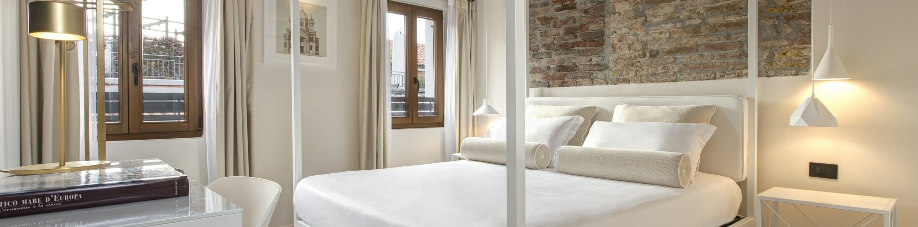 Elegantes Schlafzimmer in Weiß mit großem Bett, Bücherstapel, goldener Lampe und Fenster mit weißen Vorhängen.