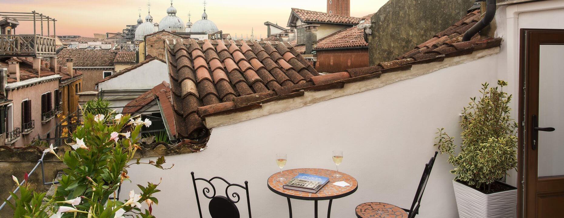 Tavolo con vino e libro su un tetto con piante fiorite.