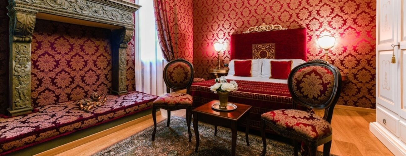 Luxurious room with red and gold wallpaper, ornate fireplace, white bed, and elegant furnishings, exuding opulence.