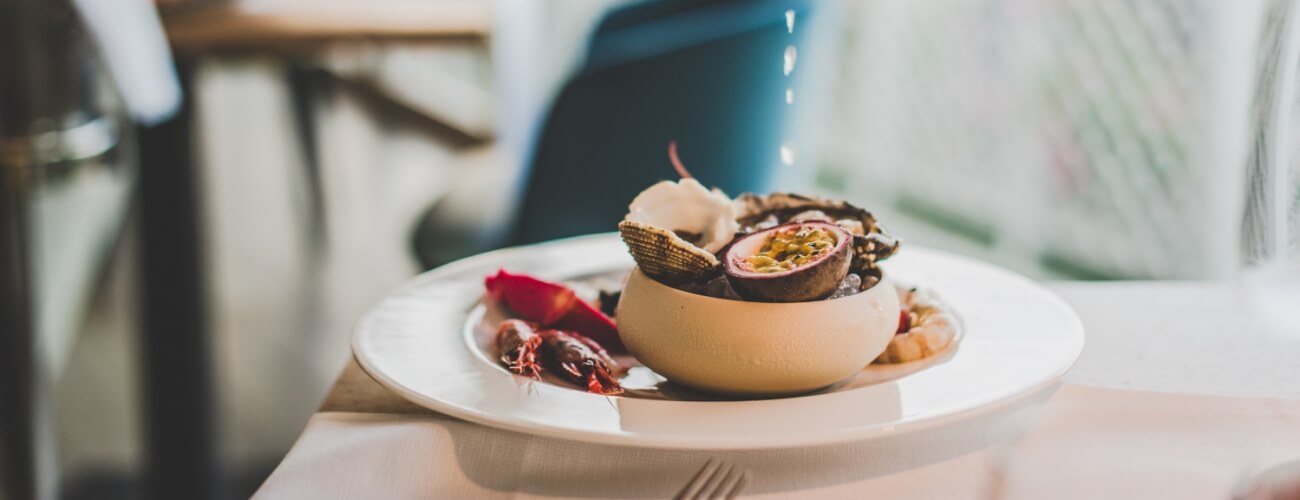 Close-up of a gourmet dish with passionfruit, corn, and red garnishes on a white plate, in a cozy café setting.
