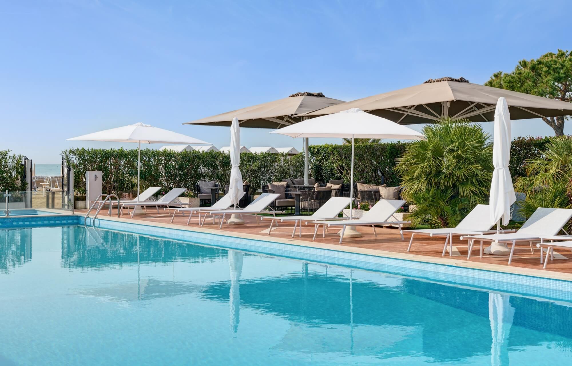 Una scena di una piscina con lettini bianchi, ombrelloni, siepi verdi e vista mare sotto un cielo blu.