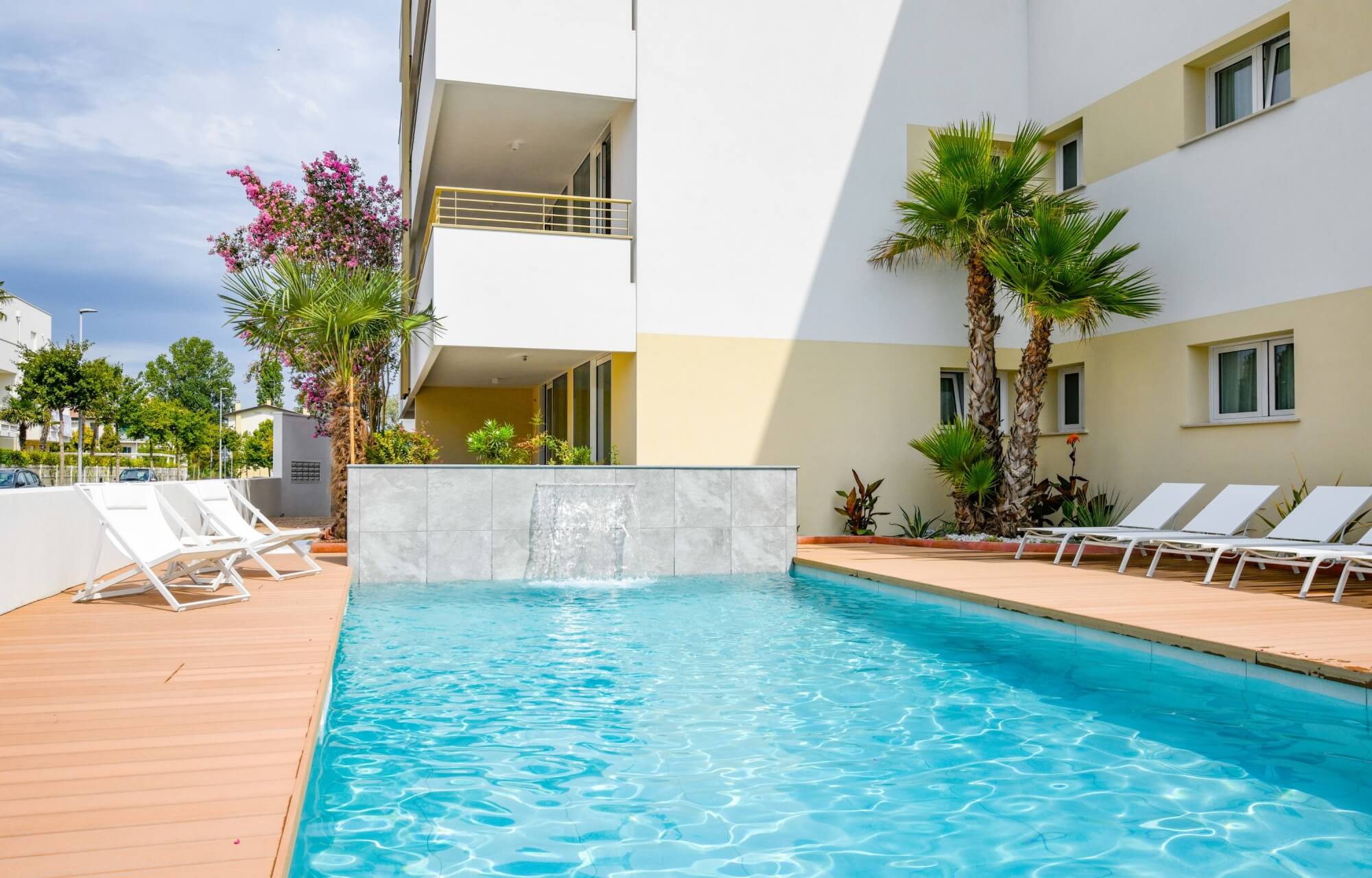 Una piscina con acqua blu, sedie a sdraio bianche, palme e un edificio bianco con accenti gialli sullo sfondo.
