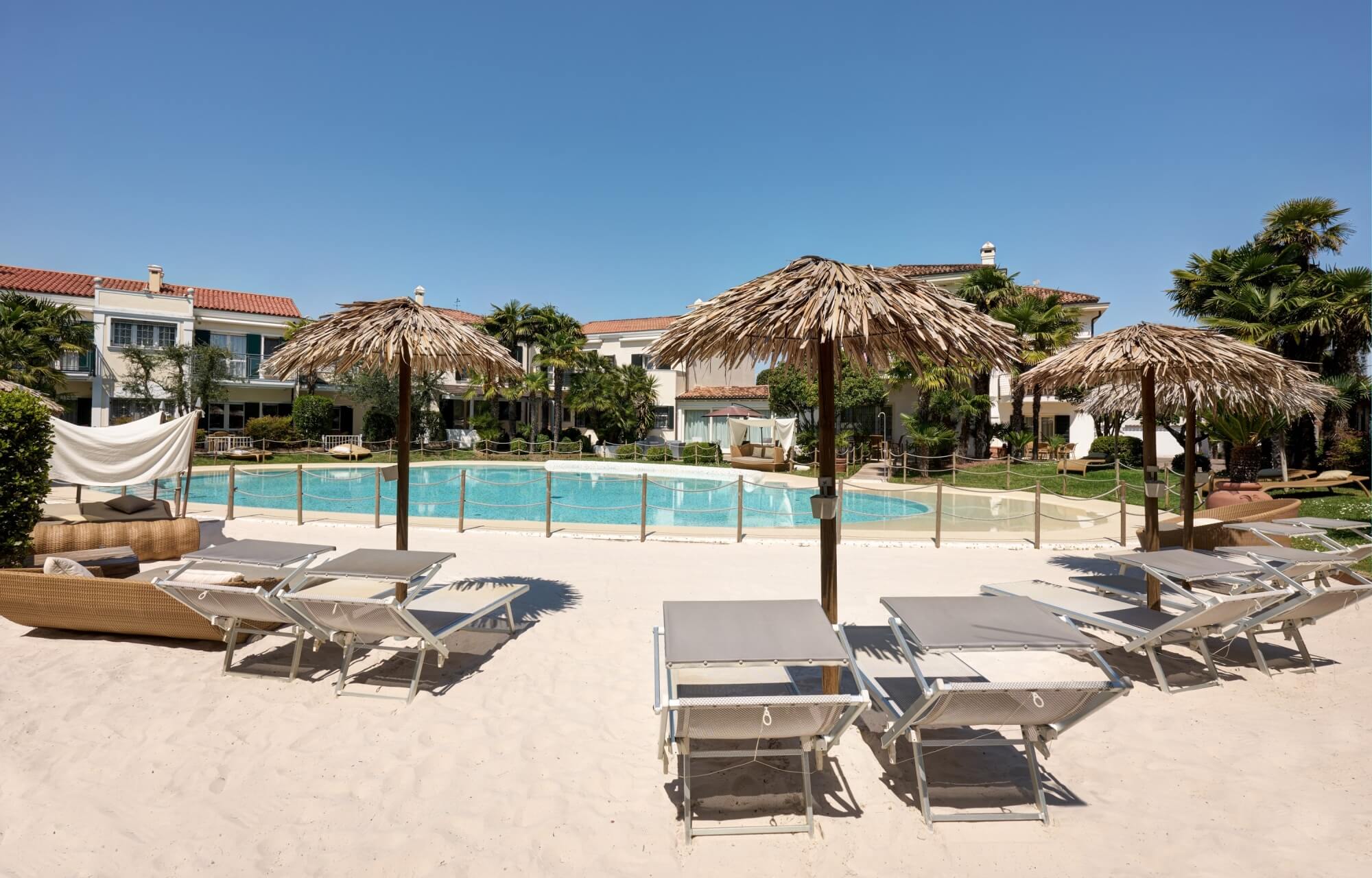 Una scena di una spiaggia con sei lettini, un piscina rettangolare e edifici sullo sfondo.