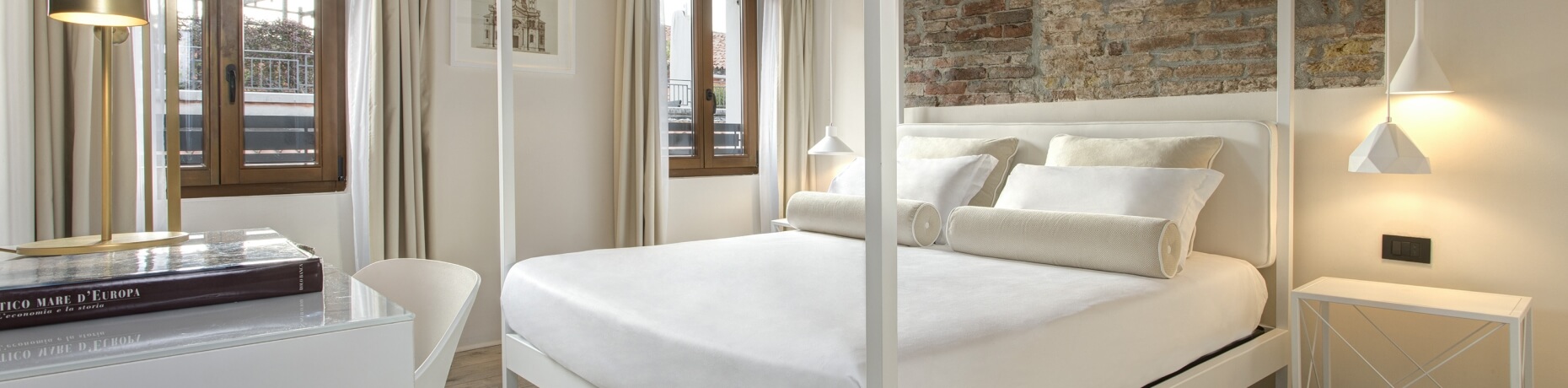 Chambre minimaliste et sereine avec des murs blancs, lit blanc, bureaux, lampes dorées et fenêtres avec rideaux blancs.