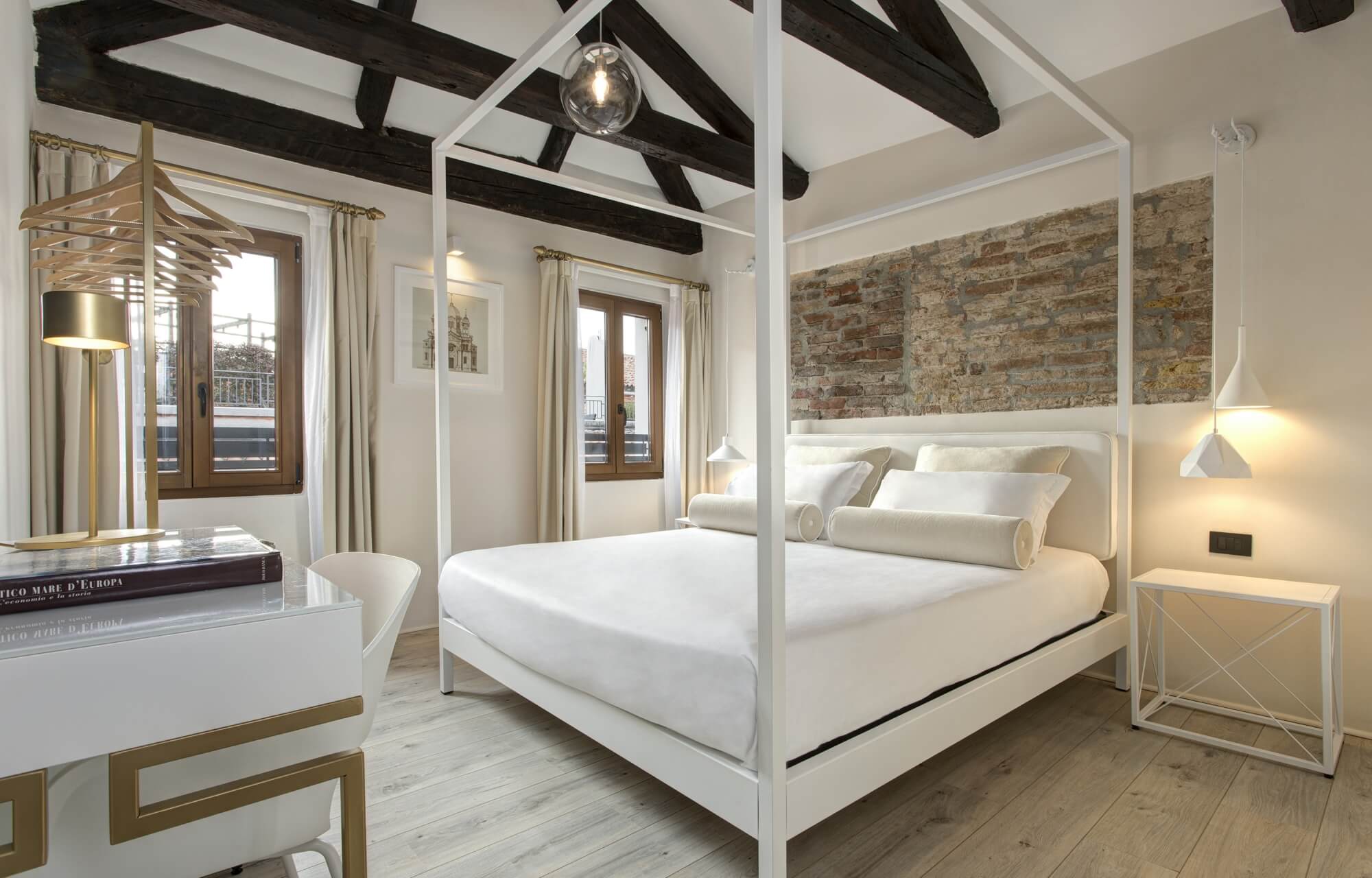 Camera elegante con letto a baldacchino bianco, scrivania, lampada e pareti bianche, illuminata da travi in legno scuro.