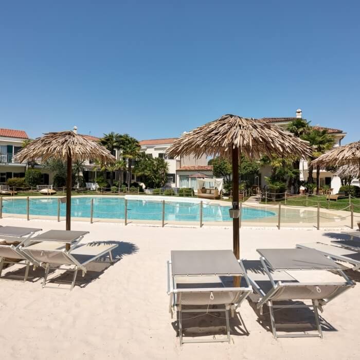 Spiaggia serena con piscina, lettini e ombrelloni, circondata da palme e edifici bianchi con tetti rossi sotto un cielo blu.