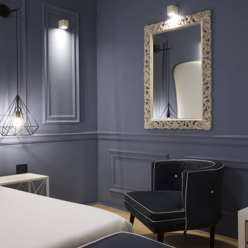 Camera d'hotel con poltrona blu, specchio bianco e letto bianco.