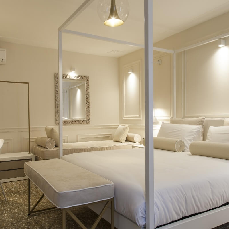 Serene bedroom with two white-framed beds, crisp linens, a bench, mirror, warm lighting, and a brown and white terrazzo floor.