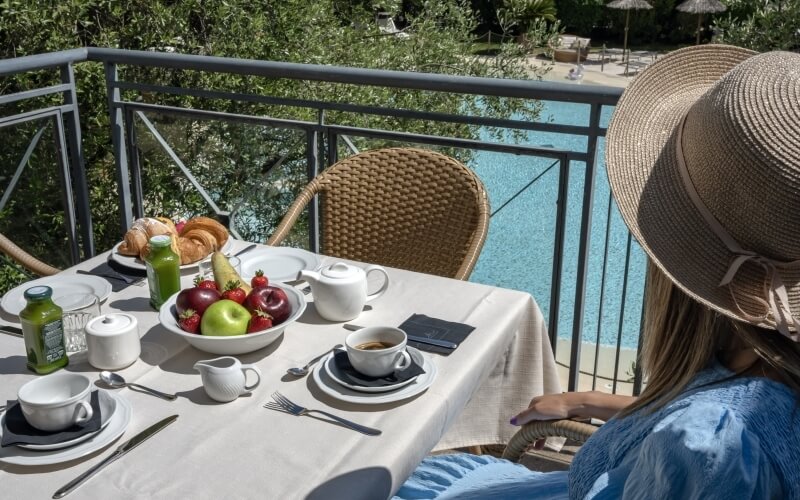 A woman in a straw hat sits at a table with fruit and drinks, overlooking a serene pool surrounded by greenery.