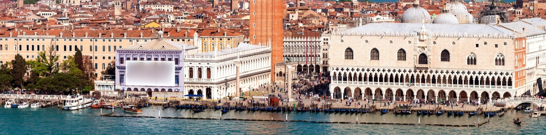 Una vivace città con un grande edificio bianco, acqua in primo piano, barche e persone che passeggiano lungo il lungomare.