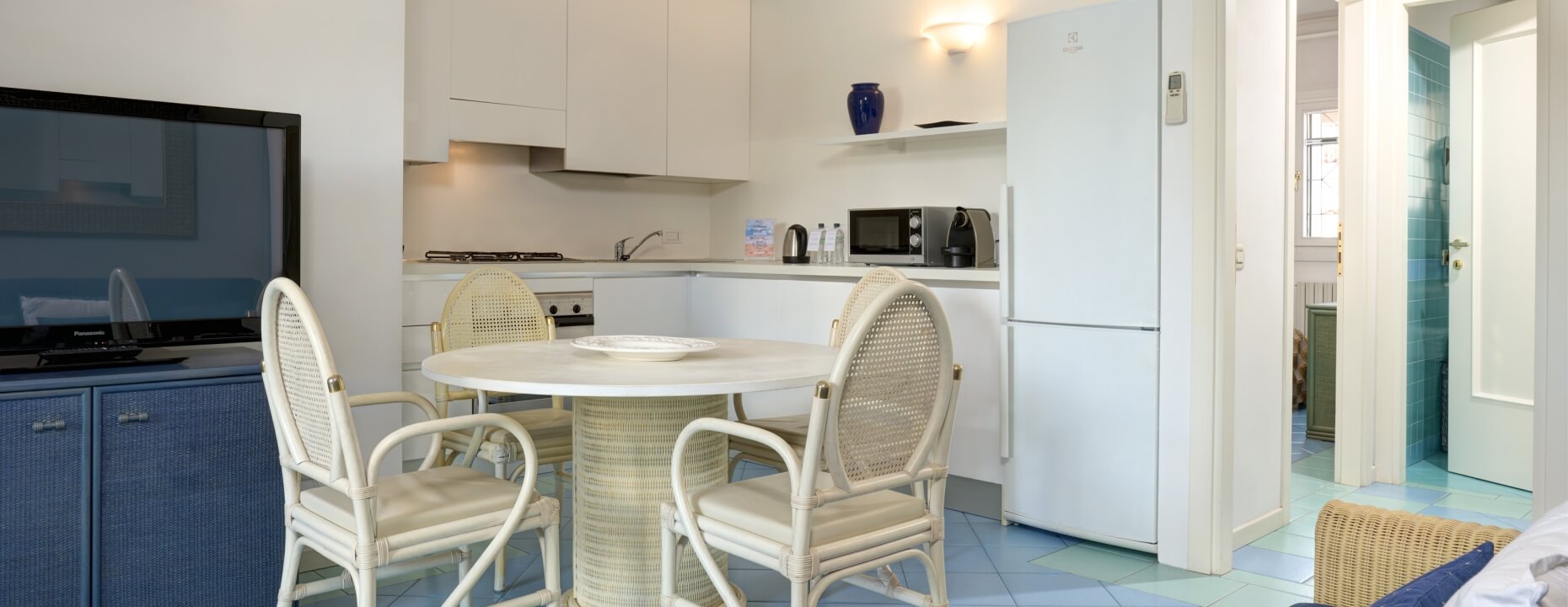Appartamento con TV, tavolo rotondo, cucina bianca e bagno turchese.