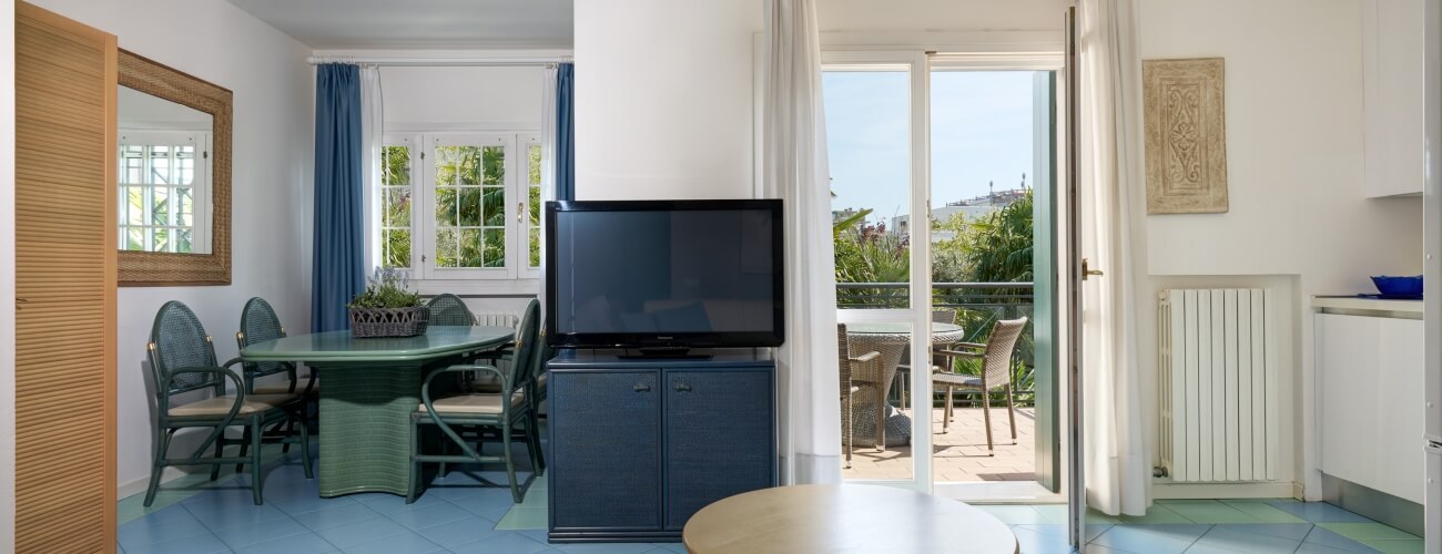 Spazio abitativo sereno con zona pranzo, piante, porte aperte su un patio e cucina adiacente con armadi bianchi.