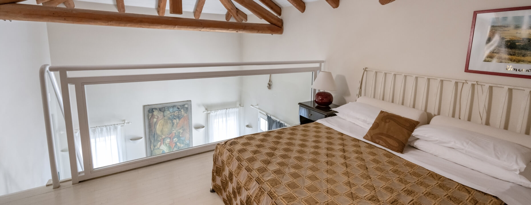 Loft bedroom with white linens, brown blanket, and wooden beams. Camera loft con lenzuola bianche, coperta marrone e travi di legno.