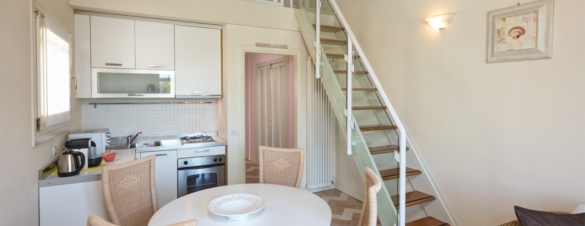 Cucina moderna con armadi bianchi, tavolo rotondo e scala in vetro.