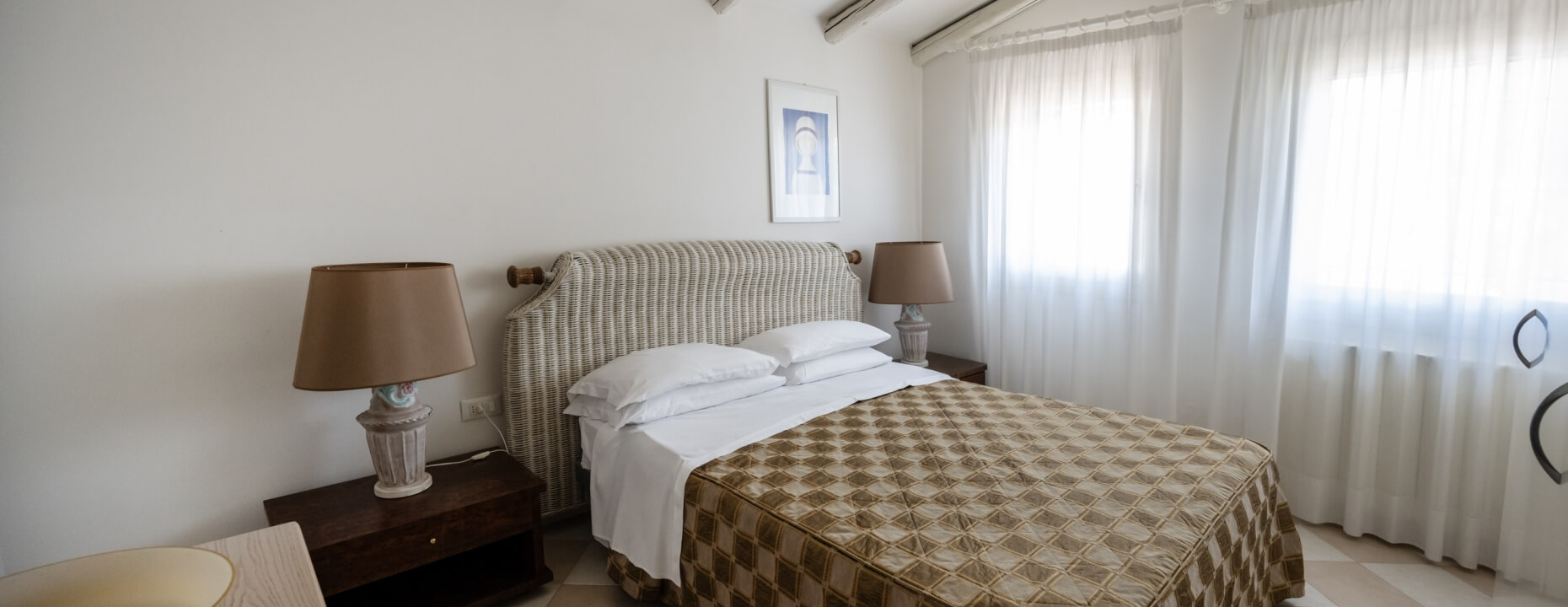 Camera d'hotel con letto, testiera in vimini e tende bianche.