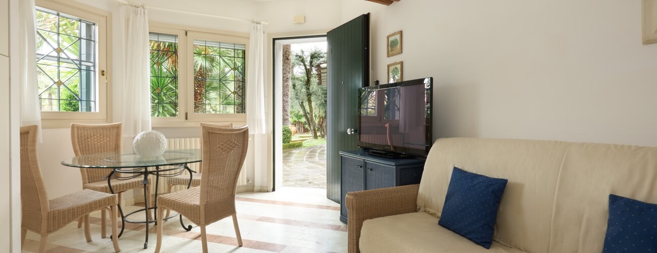 Salon serein avec canapé blanc, table en verre, chaises en osier, télévision, lumière naturelle et accès à un patio.